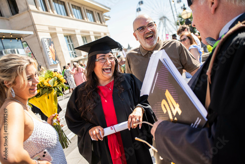 A jubilant graduate holding a diploma laughs with joy surrounded by family and a professor during an outdoor graduation ceremony, Bournemouth, Dorset UK