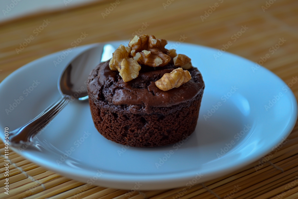 A photo of a delicious chocolate cupcake with walnuts served on a white plate with a silver spoon placed on a bamboo mat background for an invitation presentation. A photo of a delicious chocolate cupcake with walnuts served on a white plate with a silver spoon placed on a bamboo mat background for an invitation presentation.