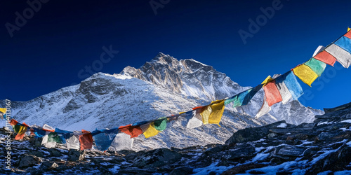 A dramatic view of a Himalayan peak at dawn, with snow glittering in the first light, prayer flags fluttering in the breeze, and a deep blue sky. The awe-inspiring and sacred scene feels timeless. photo