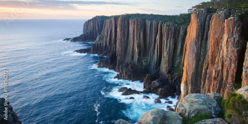 A dramatic coastal vista with cliffs rising steeply from turquoise waters, waves crashing against the rocks, and the horizon glowing with the light of the setting sun. The majestic and untamed setting