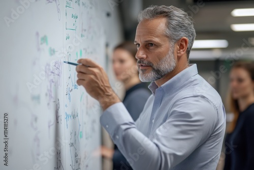 A focused professional explains concepts on a whiteboard, emphasizing collaboration in a modern workspace with engaged colleagues observing behind him. photo