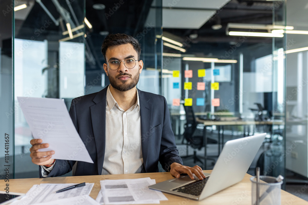 Businessman in formal attire holding documents by a laptop, showcasing confidence in a modern office. Ideal for concepts of entrepreneurship, corporate focus, and professional meetings. Businessman in formal attire holding documents by a laptop, showcasing confidence in a modern office. Ideal for concepts of entrepreneurship, corporate focus, and professional meetings.