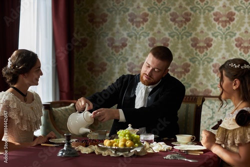 Portrait of classic young gentleman in black tailcoat pouring tea from white teapot while sitting with two women at elegantly set dining table, copy space photo