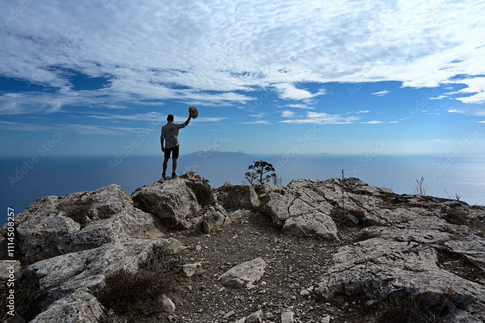 Obraz premium Die Silhouette einer Person steht auf einem Berg hoch über dem blauen Meer und winkt mit seinem Hut zu einer benachbarten Insel .