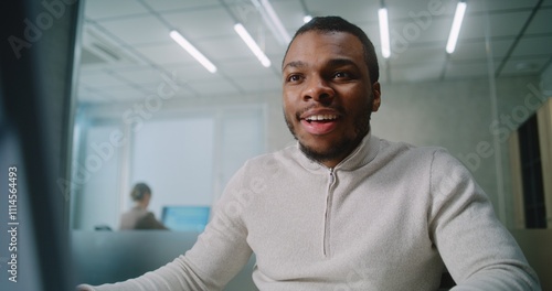 Excited young businessman rejoices and celebrates successful business deal with customer, uses computer, talks by mobile phone. African American marketing specialist works in modern office. E-commerce