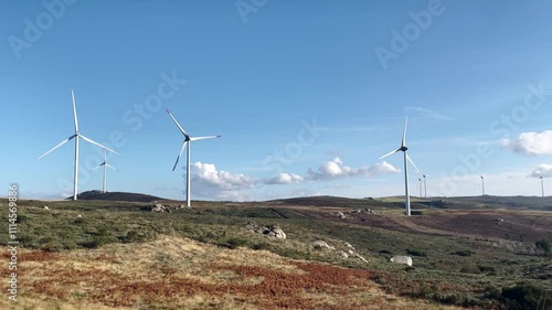 Wind turbines spinning in the countryside of Felgueiras, Portugal, with blue skies, rocks, shrubs, and clouds, showcasing the peaceful landscape