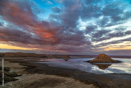 Kazakhstan, Ustyurt National Park, Karynzharyk. At dawn, 2 lonely mountains are surrounded by a thin layer of water with a perfect reflection. Clouds are illuminated by the sun from below