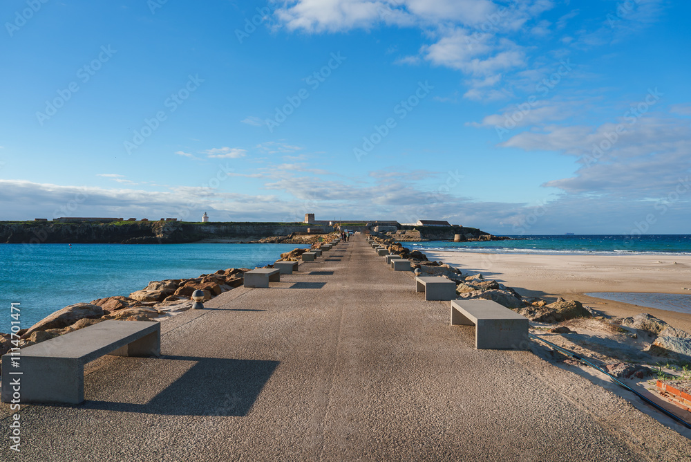 A long pier with concrete benches extends towards the Castle of Tarifa, surrounded by vibrant blue waters and a sandy beach under a clear sky. A long pier with concrete benches extends towards the Castle of Tarifa, surrounded by vibrant blue waters and a sandy beach under a clear sky.