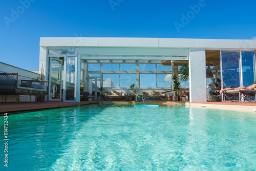 A rooftop pool area features modern Marrakech design, with sleek structures, large glass windows, seating, and lush plants under a clear blue sky.