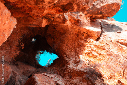 A vibrant view of a rocky cave entrance illuminated by sunlight, showcasing vivid orange and red tones with a glimpse of the bright blue sky in the background.