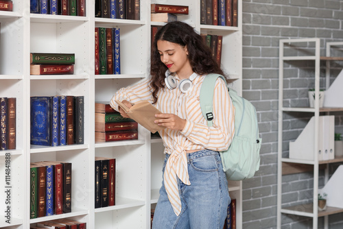 Teenage girl with backpack and headphones reading a book in a library or book...