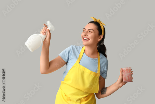 Beautiful young happy woman with sponge and bottle of detergent on grey background photo