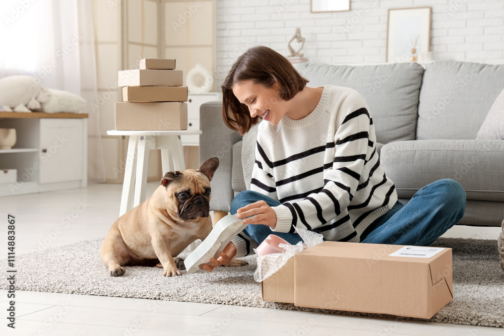 Fototapeta premium Pretty young woman with French bulldog unpacking dog bowl from parcel at home