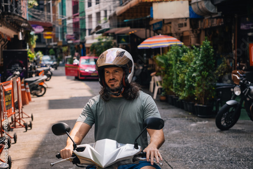 Horizontal portrait of mid adult man driving a scooter with an open face helmet in the southeast of Asia; front view in a street of Sukhumvit, Bangkok, Thailand photo