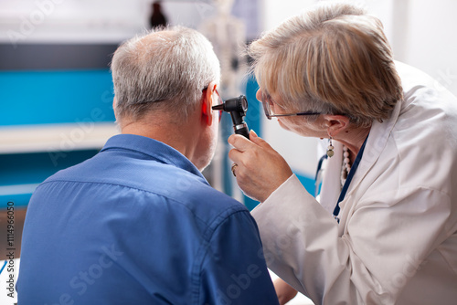 Male pensioner patient in hospital room has his ear examined by skilled senior doctor. Retired female physician using otoscope on old white man, ensuring professional diagnosis in medical office. photo