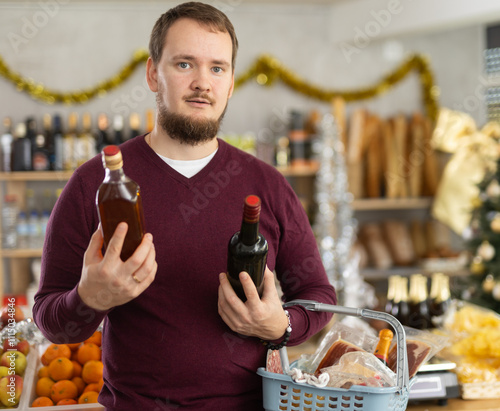 Pensive bearded young man in casual sweater, surrounded by Christmas decorations, selecting festive snacks and drinks in local grocery store, comparing bottles of whiskey and pondering over choice.. photo