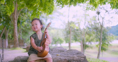 Happy smiling Joyful Asian child girl sitting on a rock playing a ukulele guitar Outdoors in a beautiful park with lush greenery in the background. photo