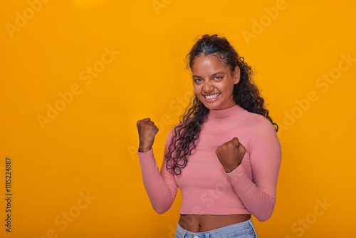 Young Indian woman stands confidently with her fists raised in triumph. Her bright smile complements the vibrant yellow background, radiating positivity and energy in a playful atmosphere.