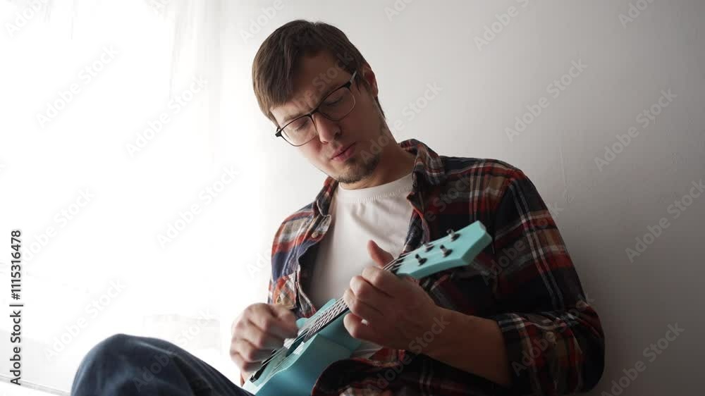 Young musician is sitting by the window, playing a blue ukulele. He is concentrating on his playing and seems to be enjoying the music Young musician is sitting by the window, playing a blue ukulele. He is concentrating on his playing and seems to be enjoying the music