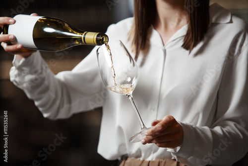 Professional woman sommelier pours alcoholic beverage from bottle into wine glass standing in cellar on shelf background.  photo