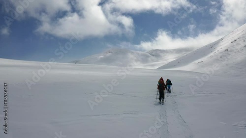 Two skiers traverse a vast snow-covered valley surrounded by majestic mountain peaks under a bright blue sky with scattered clouds. A breathtaking winter adventure in pristine nature
