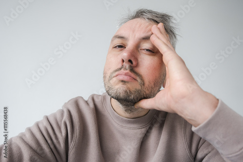 Close-up portrait of a tired or stressed man, hand to his face. His expression conveys weariness, fatigue, or contemplation. The neutral background emphasizes his emotional state