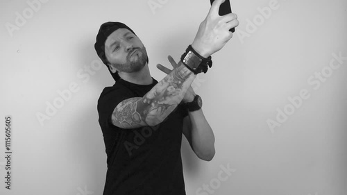 Grayscale footage of a tattooed man posing to a smartphone or phone camera and taking a selfie while showing a peace sign with his other hand. Man looks smug and happy to post on social media.