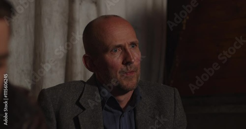A Caucasian man in a dark cabin looks around and talks to people sitting around him