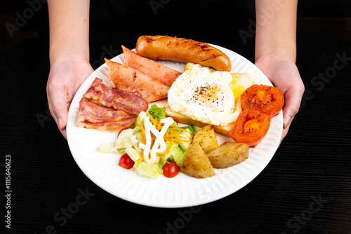Hand holding American breakfast set with eggs,bacon,toast, potatoes, tomatoes, mayonnaise sauce and sausage on black background.