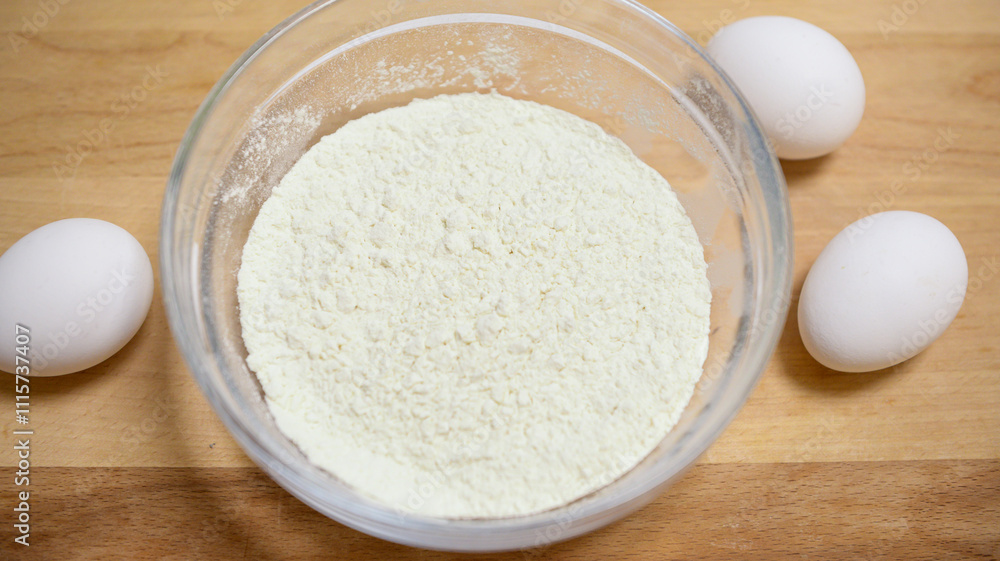 a bowl of flour stands on a wooden surface, three white chicken eggs lie nearby. cooking at home, homemade baking, following a recipe, preparing ingredients, kitchen tools. a bowl of flour stands on a wooden surface, three white chicken eggs lie nearby. cooking at home, homemade baking, following a recipe, preparing ingredients, kitchen tools.