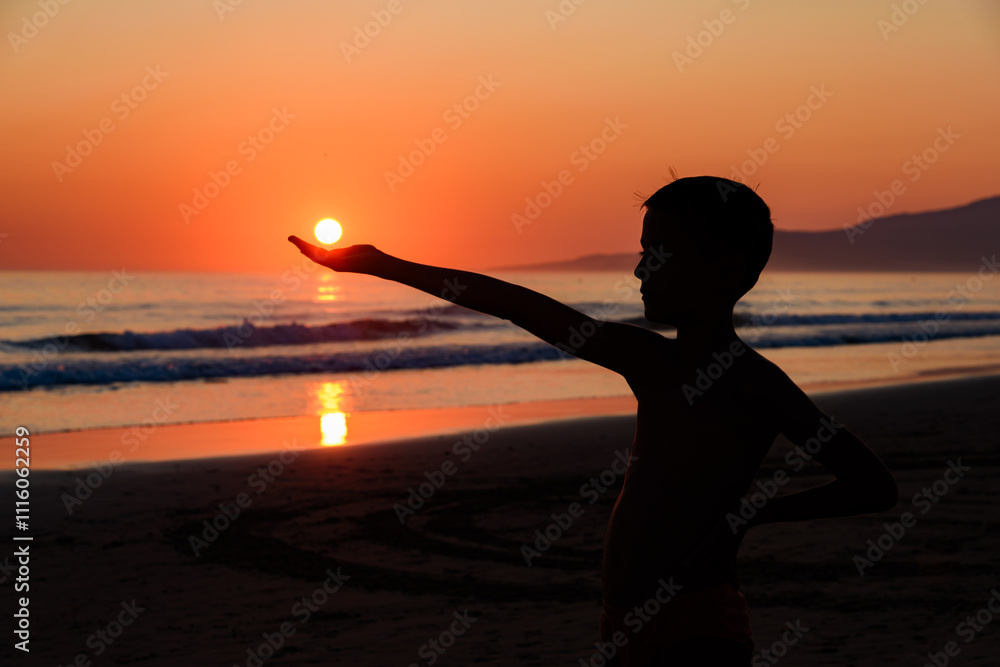 Boy on the beach with sunset. The boy is holding the sun in his hand. Silhouette of a hand holding the sun, in front of the sea at sunset.