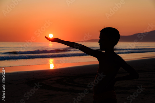 Obraz na płótnie Boy on the beach with sunset. The boy is holding the sun in his hand. Silhouette of a hand holding the sun, in front of the sea at sunset. w salonie