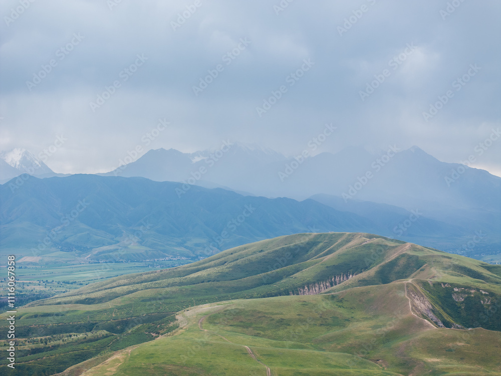 Fototapeta premium Drone view of the mountains in cloudy weather. Large dark clouds in the sky. Green and yellow grass in a field on the hills.