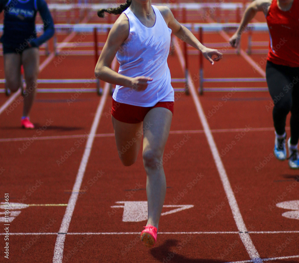 Female Athlete Competing in a Hurdle Race on a Red Track During a Sunny Day at a Sports Event Female Athlete Competing in a Hurdle Race on a Red Track During a Sunny Day at a Sports Event