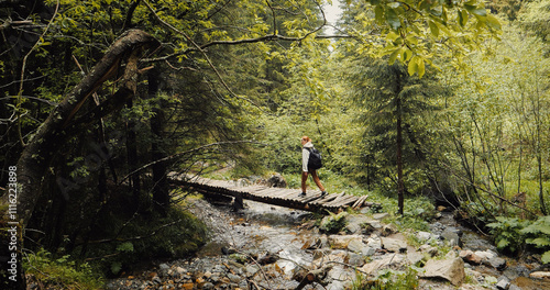Tourist caucasian girl walking on a rocky hiking path along a river with bridge in dence forest. Back view of woman lost in the woods by the river. Active holiday concept