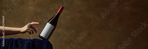 Mockup showing red wine bottle with blank label, being gracefully balanced by hand adorned with decorative red-stoned ring, against rich brown textured background. Concept of alcohol beverage.