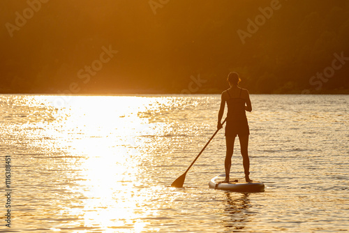 Back view of slim sporty young female standing on sup board with sunset reflected on calm water on hot summer evening; golden hazy sundown with lady on stand up paddleboard; beautiful summer evening .