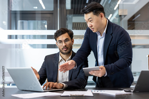 Two business professionals engage in a collaborative discussion in a modern office environment. One stands holding a tablet, while the other works on a laptop. The setting highlights teamwork. photo