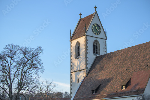 Gotischer Kirchturm einer Dorfkirche mit Kirchturmuhr und Schattenwurf eines kahlen Baumes vor blauem Himmel photo