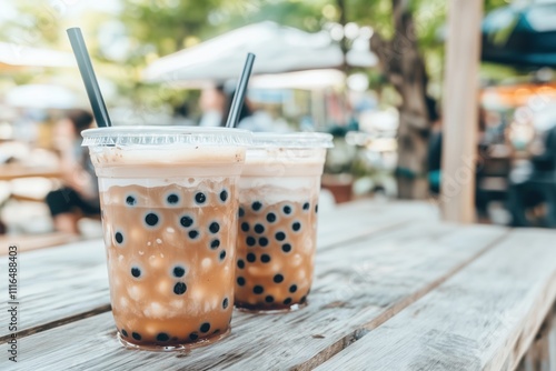 Two Cups of Refreshment: A close-up shot of two iced bubble tea drinks with tapioca pearls, resting on a wooden table with a blurred background of a bustling outdoor cafe.