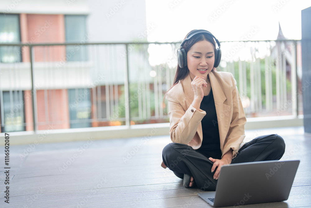 Fototapeta premium Asian businesswoman wearing headphones working on laptop while sitting with legs crossed on terrace floor, enjoying fresh air and modern technology for remote work