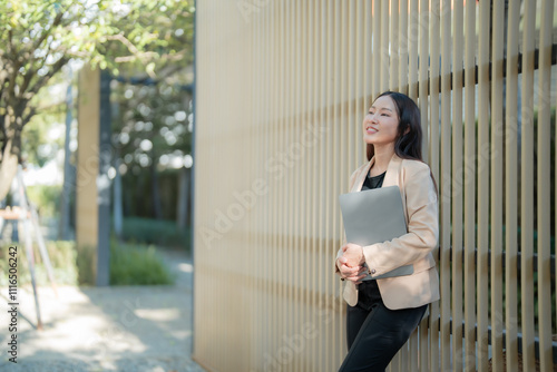 Young businesswoman is holding laptop and leaning against wooden wall in a park, enjoying break time and fresh air, looking away and smiling Free Download Young businesswoman is holding laptop and leaning against wooden wall in a park, enjoying break time and fresh air, looking away and smiling - GFXLoad