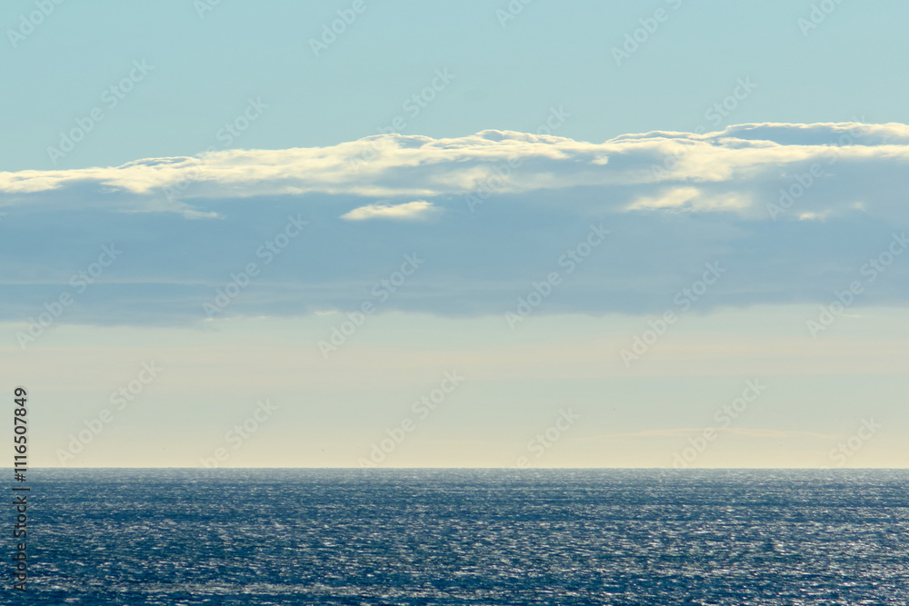 Fototapeta premium An expansive sea view with sunlight reflecting on the water. A large, illuminated cloud stretches across the sky, enhancing the serene and dynamic scenery.