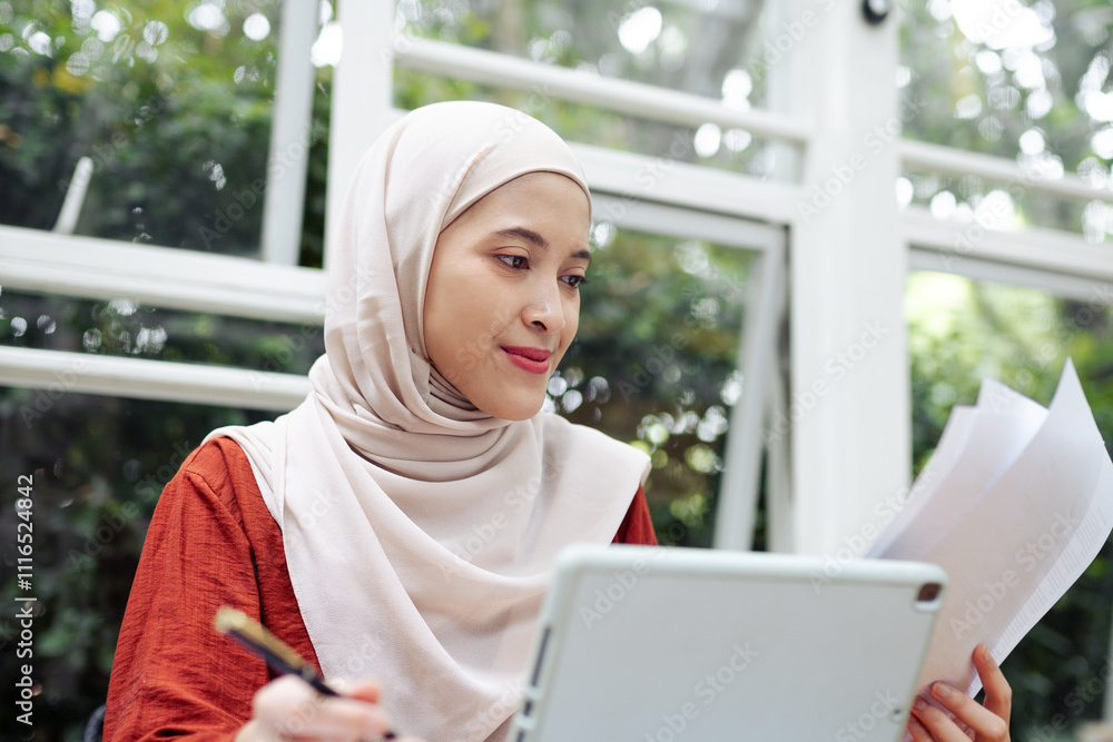A focused woman in a hijab examines documents while using a digital tablet indoors, demonstrating multitasking and professionalism in her work environment. Freelance work concept A focused woman in a hijab examines documents while using a digital tablet indoors, demonstrating multitasking and professionalism in her work environment. Freelance work concept