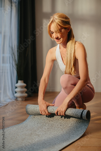 A blonde woman rolls up her yoga mat after a workout, with sunlight streaming through a large window.