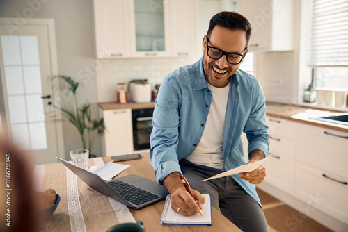 Happy man taking notes while analyzing his home finances in kitchen. photo