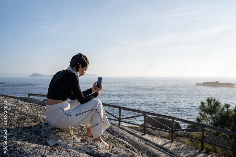 Young woman relaxes on a coastal rock, soaking in the sunshine and ocean view while using her smartphone Young woman relaxes on a coastal rock, soaking in the sunshine and ocean view while using her smartphone