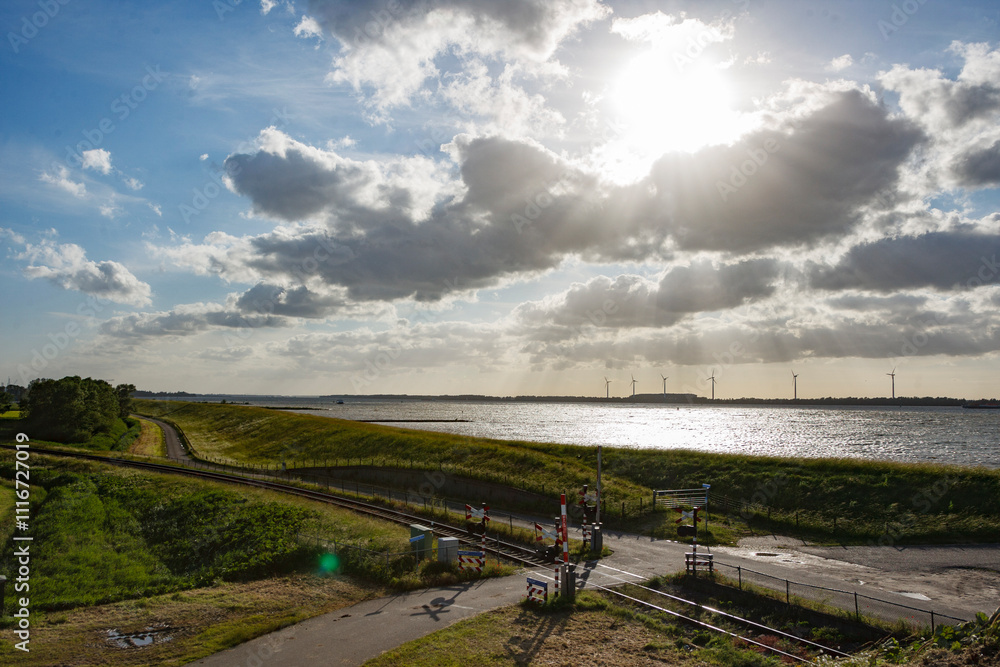 Fototapeta premium Beautiful landscape with sun shining over a river and wind turbines under a cloudy sky in the countryside