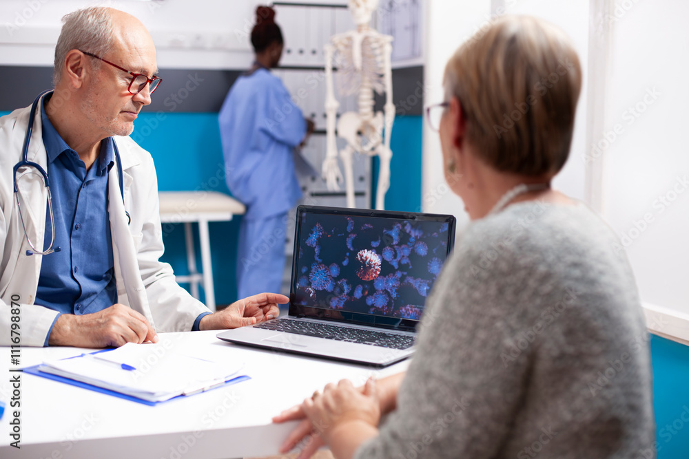 Fototapeta premium Senior male physician in lab coat discusses virus image on laptop with retired woman in well equipped hospital room. Old doctor and female patient looking at enlarged viral cells on digital device.