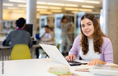 Young interested woman sitting at table in library, using laptop and books for research. Self-education concept..